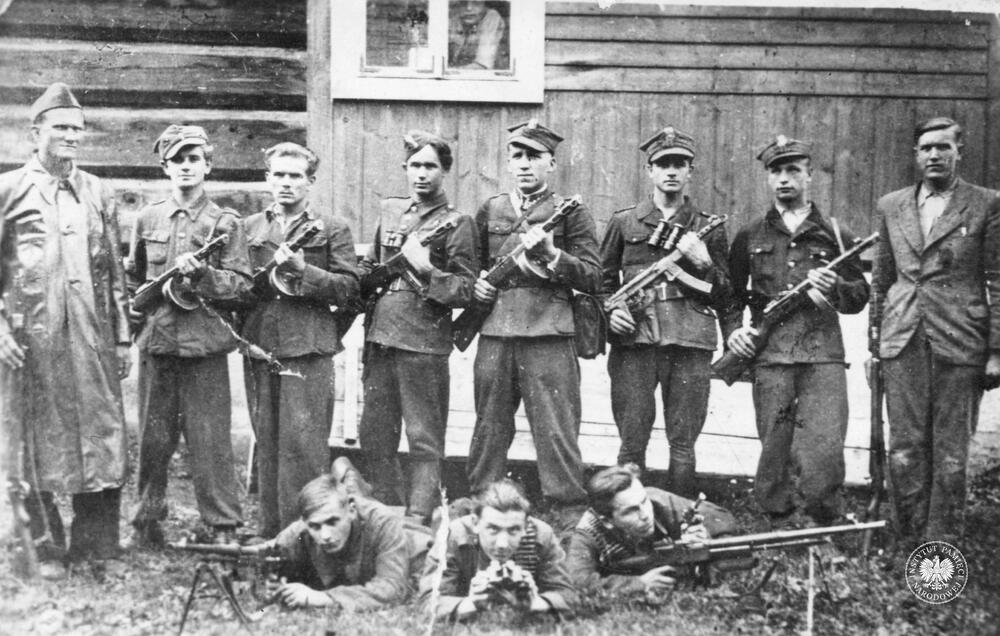 Grupa jedenastu partyzantrów z oddziału Harnaś pozuje do zdjęcia trzymając broń przed nieznanym budynkiem w miejscowości Toporzysko, fot AIPN, sygn. IPNKr-3-3-18-1-1