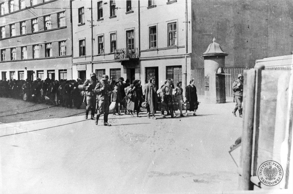 Wysiedlenie Żydów z getta krakowskiego do obozu płaszowskiego. Żydowska ludność Krakowa, niosąc swój dobytek, pod eskortą SS-manów opuszcza teren getta. Na ostatnim planie kamienice stojące przy ulicy Lwowskiej. Marzec 1943 r. (fot. z zasobu IPN, sygn. IPN Kr 1/9023)