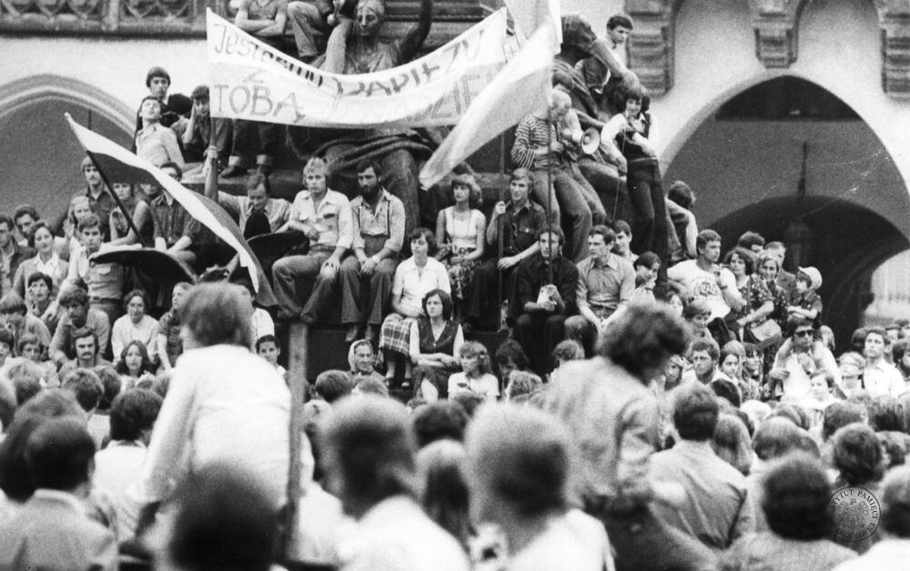 Grupa ludzi na cokole pomnika Adama Mickiewicza w dniu przybycia do kościoła Mariackiego papieża Jana Pawła II. Widiczny transparent z hasłem: „Jesteśmy z Tobą Papieżu nadziei”. Na ostatnim planie Sukiennice, 9 czerwca 1979 r., z zasobu AIPN, sygn. IPNKr-7-7-1-140-1