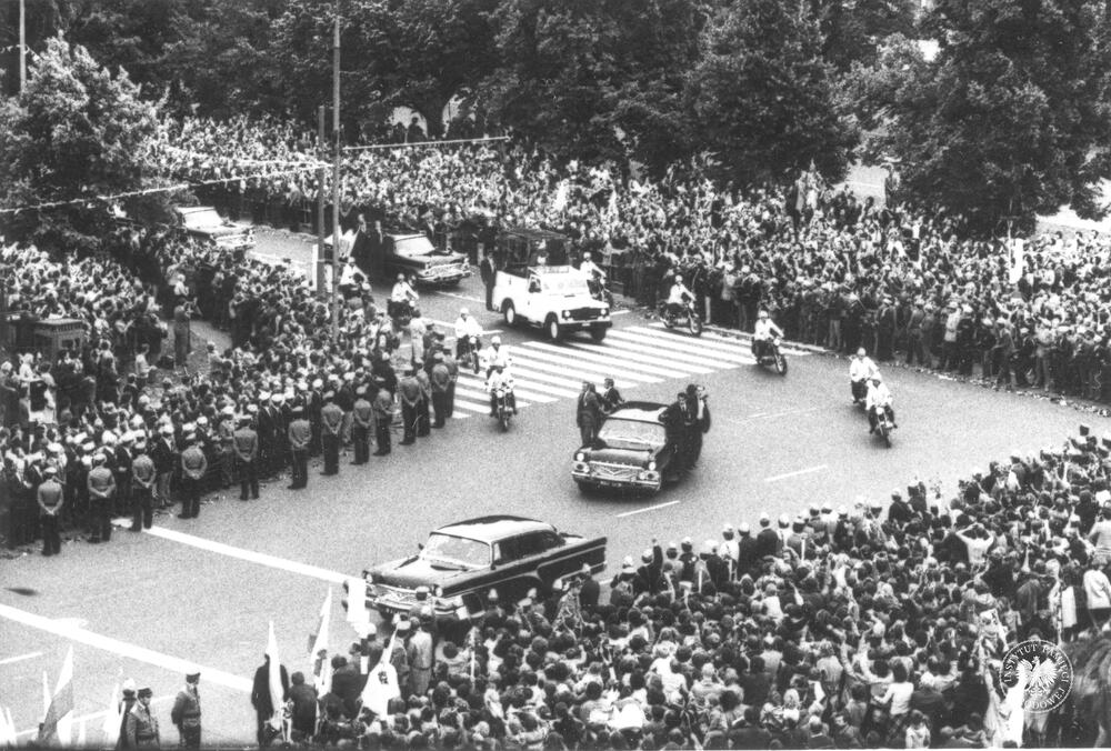 Druga pielgrzymka Jana Pawła II do Ojczyzny. Kolumna samochodów papieskich, Jan Paweł II widoczny w papamobile w asyście milicji na motocyklach, przejeżdża wśród tłumów rozstawionych po obu stronach ulicy na skrzyżowaniu ulic Wawelskiej i Żwirki i Wigury przed pomnikiem Lotnika. z zasobu AIPN, sygn. IPNBU-7-14-10-55-1