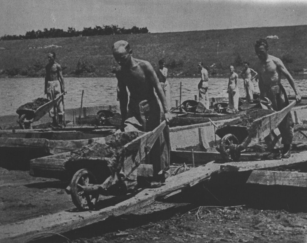 Robotnicy Baudienstu w czasie prac budowlanych nad brzegiem Wisły. Na pierwszym planie trzech młodych mężczyzn rozebranych do pasa, pcha taczki po ułożonych na placu budowy deskach. W głębi kilku robotników stoi i przygląda się pracującym. Pod obrazem napis w języku niemieckim: "BAUDIENST BEIM GALEERENBETRIEB AN DER WEICHSEL BEI KRAKAU" (Praca robotników Baudienstu nad brzegiem Wisły w Krakowie). Fot: Domena Publiczna, Źródło: IPN, Sygnatura: IPNKr-2-15-12-2