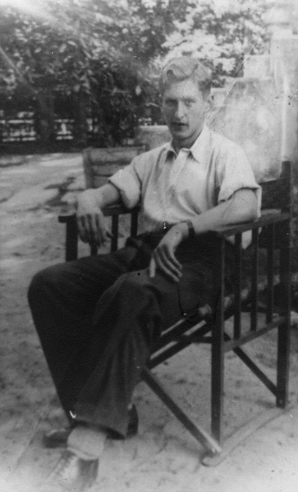 Andrzej Romocki pseudonim „Andrzej Morro" w jasnej koszuli, ciemnych spodniach siedzący na krześle w ogrodzie. W tle schody do domu, ogrodzenie, drzewa. Fot: Domena Publiczna, Źródło: AIPN, Sygnatura: IPNBU-13-1-55-2