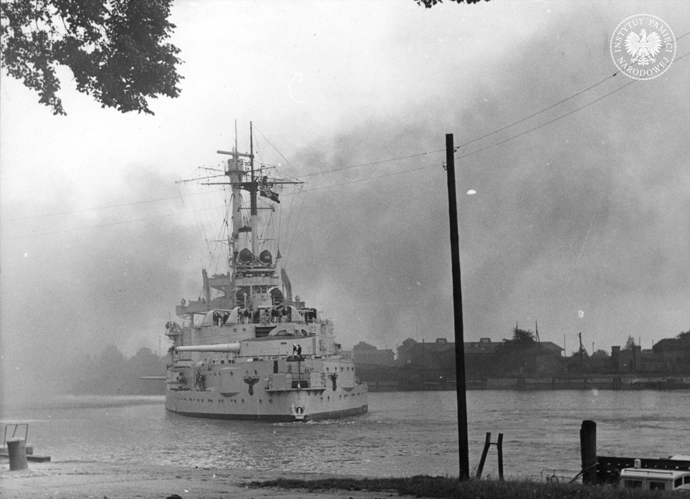 Niemiecki pancernik szkolny „Schleswig-Holstein” ostrzeliwuje polską placówkę (Wojskową Składnicę Tranzytową) na Westerplatte w Gdańsku, stojąc w poprzek kanału portowego. Autor: Hans Sönnke. Gdańsk, 1 IX 1939 r. (fot. z zasobu IPN)