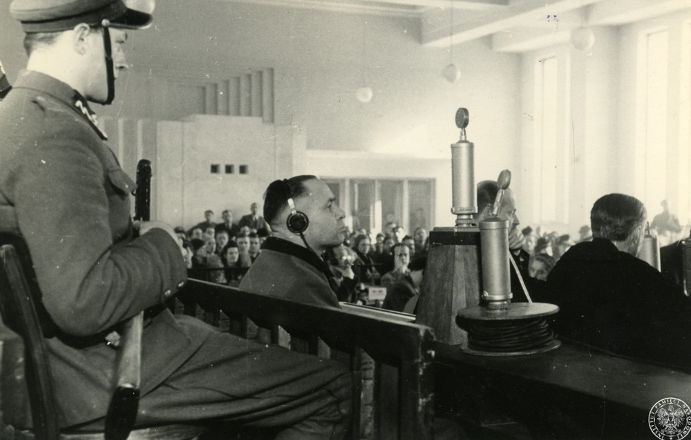 Rudolf Höss (pośrodku, w słuchawkach), były komendant KL Auschwitz-Birkenau, siedzi na ławie oskarżonych w czasie swojego procesu przed Najwyższym Trybunałem Narodowym. Po lewej jeden z siedzących za oskarżonym funkcjonariuszy milicji uzbrojony w tzw. pepeszę. Po prawej w pierwszym rzędzie siedzą m.in. obrońcy z urzędu, marzec 1947 r. Fot. AIPN