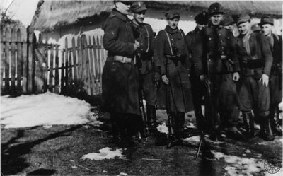 Grupa umundurowanych partyzantów z oddziału AK „Nerwy”, 1943–1944. W tle widoczna chałupa wiejska kryta strzechą. Fot. z zasobu IPN