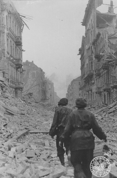 Zniszczone w czasie walk budynki przy jednej z ulic. Na pierwszym planie grupa powstańców idąca szeregiem wśród gruzów. 1944 r. (AIPN)