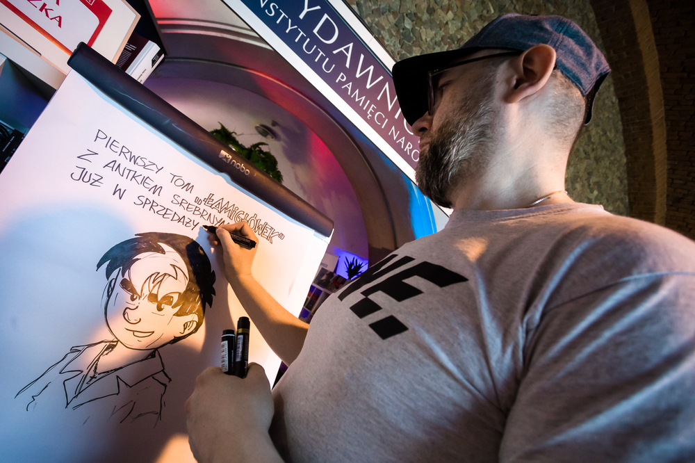 Hubert Ronek, twórca komiksowej serii IPN „Wojenna odyseja Antka Srebrnego” oraz „Aga, Adam i Hamlet na tropie tajemnic” rysuje i podpisuje – Targi Wydawców Katolickich w Warszawie, 23 kwietnia 2023. Fot. Sławek Kasper.