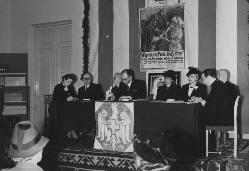 Prezydium Walnego Zjazdu Delegatów i Kół Polskiego Białego Krzyża w Kasynie Garnizonowym w Warszawie, 2 grudnia 1938 r. W środku przewodniczący Zjazdu prokurator Turski. Za członkami Prezydium plakat z hasłem wzywającym do wsparcia Polskiego Białego Krzyża. Fot. NAC