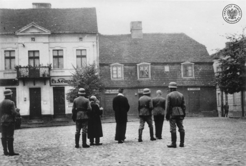 Konin, 22 września 1939 r. Dwaj skazani: Polak Aleksander Kurowski i Żyd Mordechaj Słodki, odwróceni tyłem, prowadzeni przez żołnierzy niemieckich na miejsce egzekucji na Placu Wolności w Koninie. Na skraju po prawej fragment budynku Gimnazjum Męskiego (tzw. Dom Zemełki). Fot. z zasobu IPN