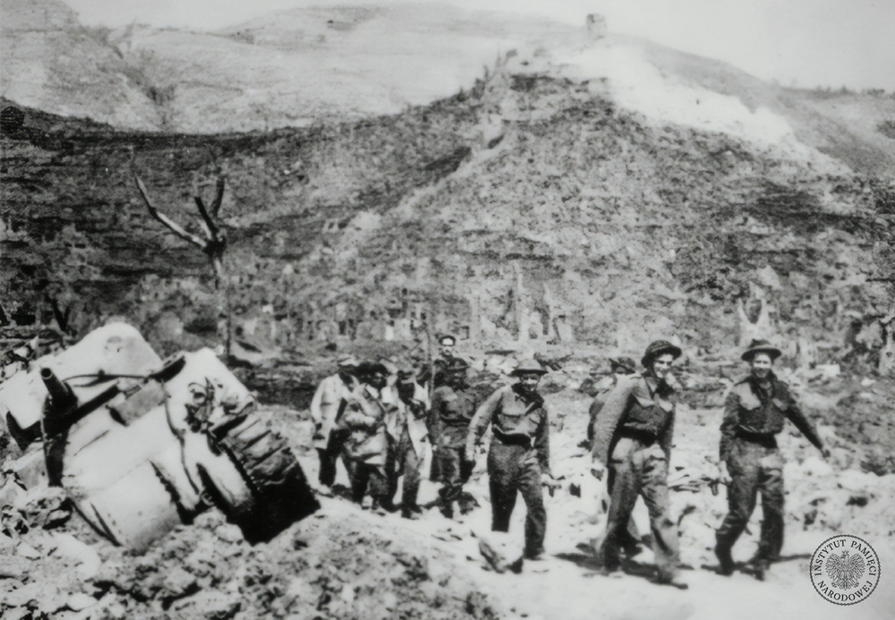 Polscy żołnierze niosą i prowadzą rannych. Na drugim planie klasztor na Monte Cassino. Po lewej zniszczony czołg M4 Sherman. Fotografia zamieszczona w pierwszym wydaniu książki Melchiora Wańkowicza Bitwa o Monte Cassino. Pod zdjęciem podpis: Znosili z gór swych rannych kolegów i drogo płacili krwią. Fot. AIPN